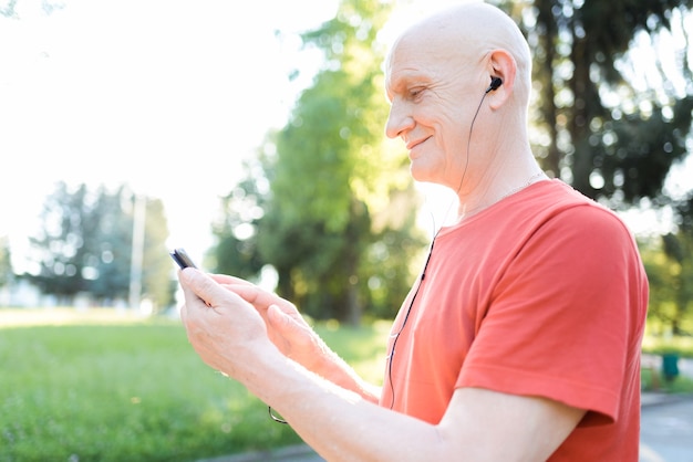 Sonriente anciano en ropa casual y auriculares blancos escuchando música desde su teléfono inteligente en el parque de la ciudad en día soleado.