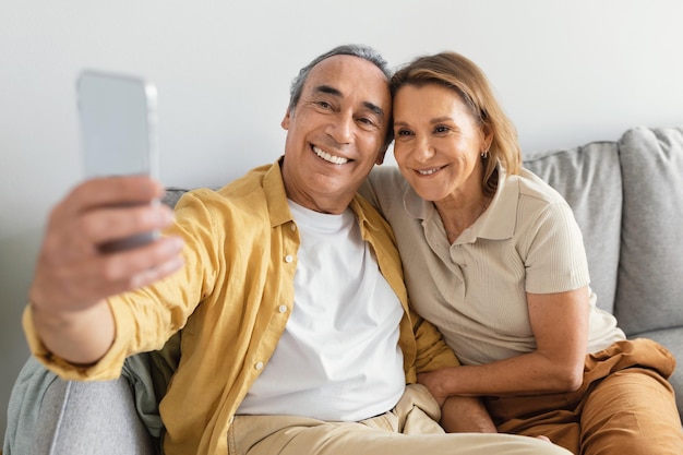 Sonriente anciano y señora tomando selfie en smartphone descansando en el sofá en casa y sonriendo a