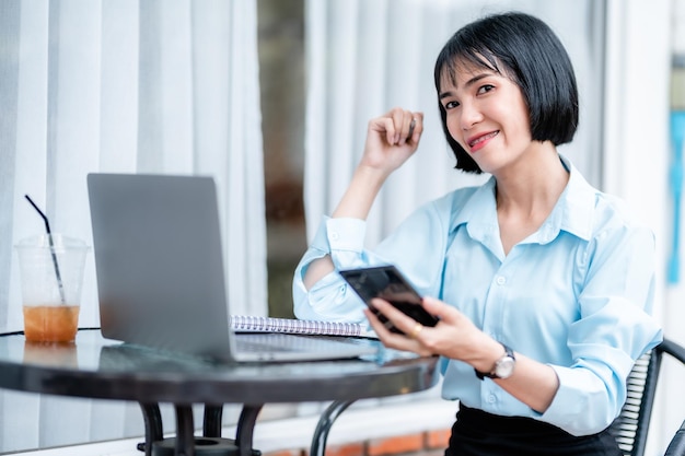 Sonriente con aparatos dentales asiáticos independientes Empresaria escribiendo un mensaje en un teléfono inteligente informal trabajando con una computadora portátil con una taza de café en el caféEstilo de vida empresarial