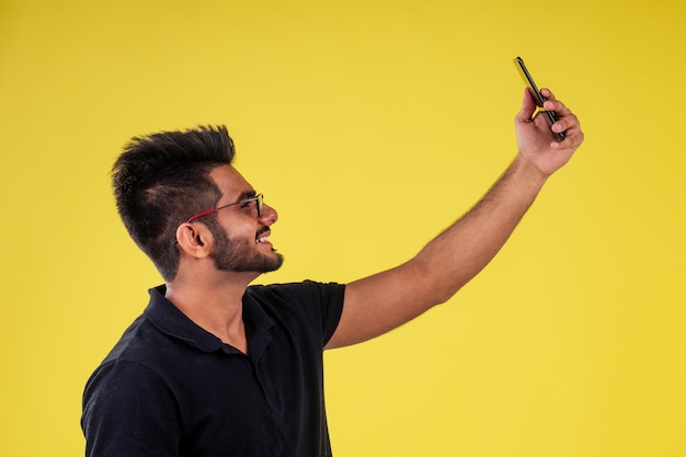 Sonriente apuesto hombre indio fotografiando selfie en estudio de fondo amarillo.