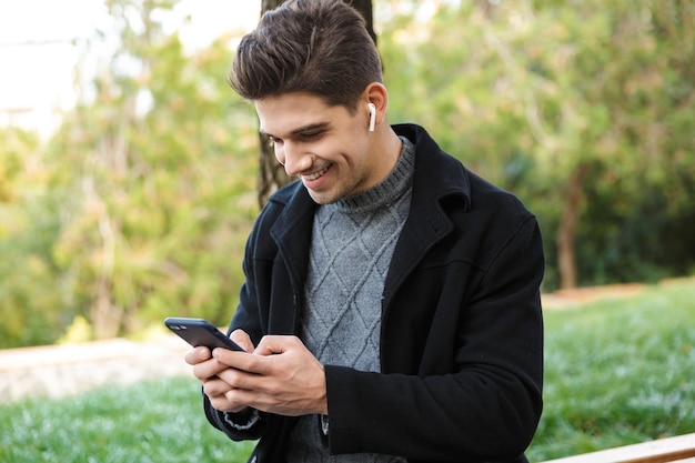 sonriente apuesto joven alegre en ropa casual caminando al aire libre en el parque verde con teléfono móvil escuchando música con auriculares.