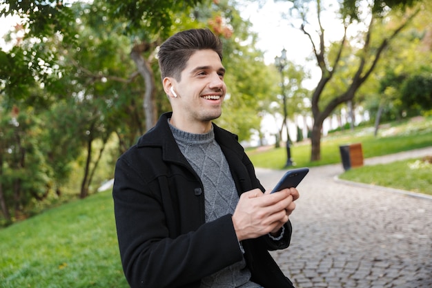 sonriente apuesto joven alegre en ropa casual caminando al aire libre en el parque verde con teléfono móvil escuchando música con auriculares.