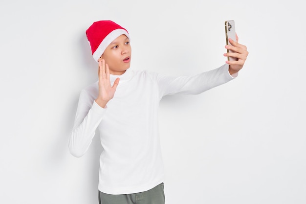 Sonriente apuesto joven asiático con camiseta blanca y sombrero de Santa Claus celebrando la Navidad en una conversación de videollamada en el teléfono móvil con fondo blanco Feliz Navidad y Feliz Año Nuevo