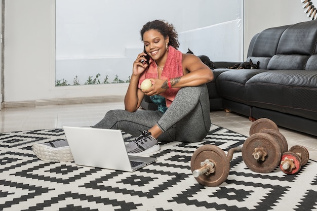 Sonriente atleta femenina negra sentada en el piso con manzana y hablando por teléfono inteligente mientras toma un descanso durante el entrenamiento en casa