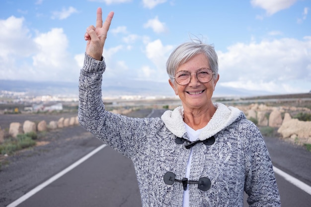 Sonriente y atractiva anciana disfrutando al aire libre mirando la cámara gesticulando victoria con la mano Anciana madura caminando sola en la montaña de la carretera vacía y el cielo azul en el fondo