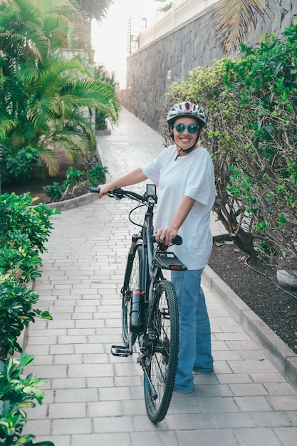 Sonriente atractiva mujer madura con casco deportivo cerca de su bicicleta eléctrica antes de correr