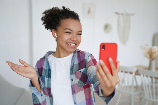 Sonriente bloguera de raza mixta en auriculares chateando por videollamada en el teléfono conduce aire en vivo en casa