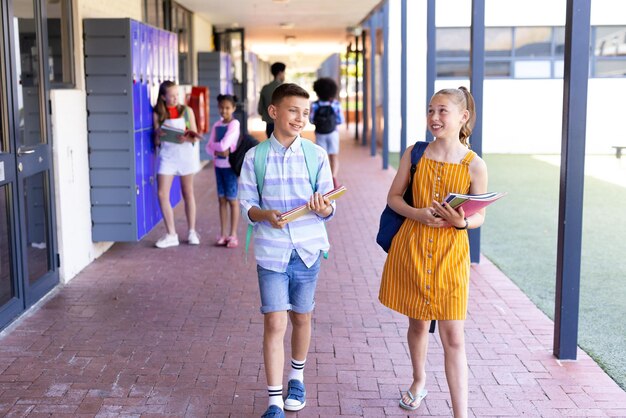 Foto sonriente caucásico de niño y niña con libros caminando por el pasillo de la escuela hablando en el espacio de copia