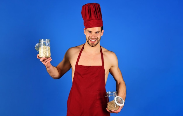 Sonriente chef masculino con sombrero y delantal burdeos sostiene frascos de vidrio con sémola y cereales profesionales