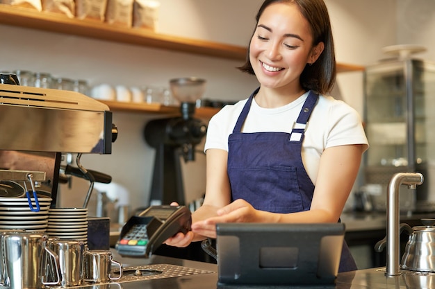 Sonriente chica asiática barista dando al cliente pos terminal ayudar a pagar con tarjeta de crédito de pie en el mostrador