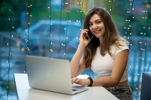 Una sonriente chica independiente que trabaja en una computadora portátil en un café y hablando por teléfono, la jornada laboral de una joven o estudiante libre