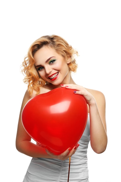 Sonriente chica rubia atractiva sosteniendo globo de corazón rojo aislado en blanco