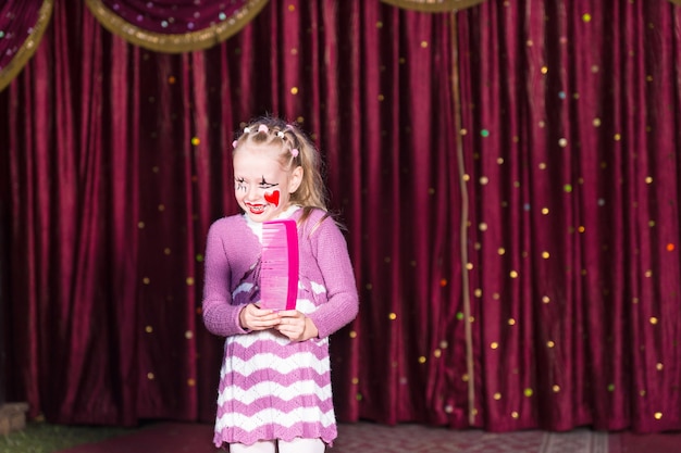 Sonriente chica rubia con maquillaje de payaso y vestido de rayas de pie en el escenario sosteniendo un peine rosa de tamaño delante de la cortina roja