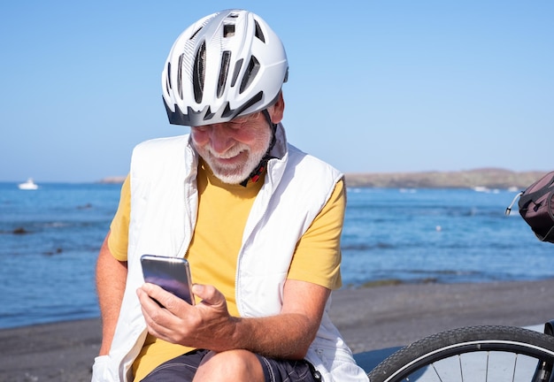 Sonriente ciclista senior barbudo con casco sentado en la playa con bicicleta eléctrica usando el teléfono