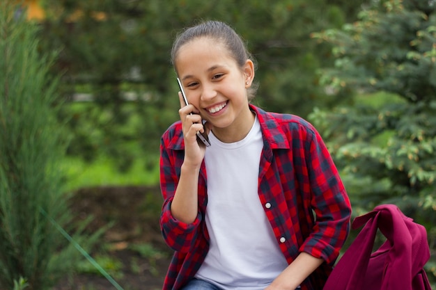 Sonriente colegiala morena se sienta en una zona del parque y se comunica en un teléfono móvil