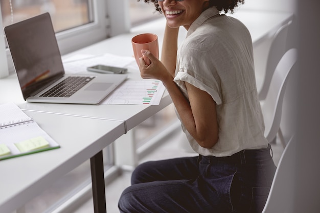 Sonriente dama afroamericana sosteniendo una taza de bebida caliente en la oficina