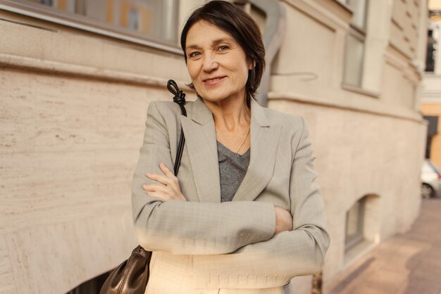 Sonriente dama caucásica de mediana edad con corte de pelo corto y oscuro mira a la cámara con los brazos cruzados frente a ella mientras está de pie en la calle Concepto de actitud positiva