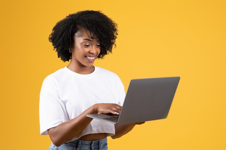 Sonriente dama negra bastante milenaria escribiendo casualmente en una computadora portátil aislada en el fondo del estudio naranja