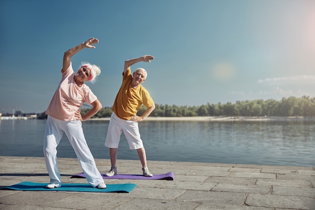 Sonriente dama senior y su cónyuge haciendo un ejercicio de flexión lateral