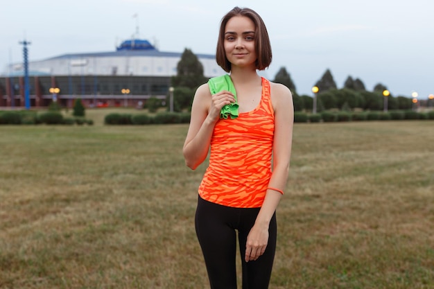 Sonriente deportista cansada en ropa deportiva naranja de pie con una toalla después del entrenamiento