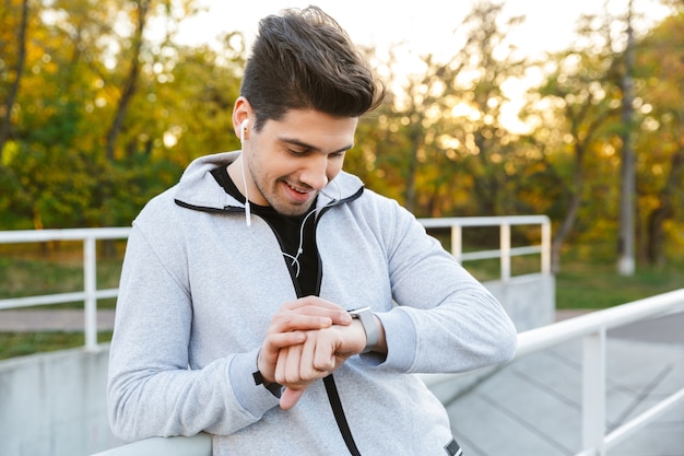 Sonriente deportista sano de pie al aire libre en la barandilla, mirando el reloj inteligente