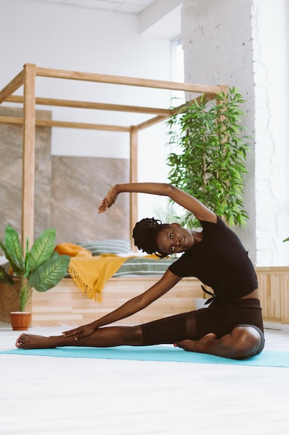 Sonriente, deportiva, flexible, descalza, piel oscura, mujer interracial estirada haciendo ejercicios de fitness de yoga en la alfombra en casa