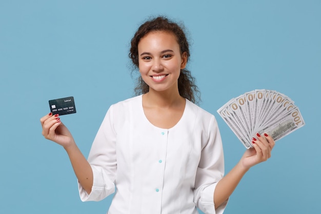 Sonriente doctora afroamericana aislada de fondo azul. Doctora en bata médica sostenga fan de dinero en efectivo en dólares, tarjeta de crédito. Concepto de medicina del personal sanitario. Simulacros de espacio de copia.