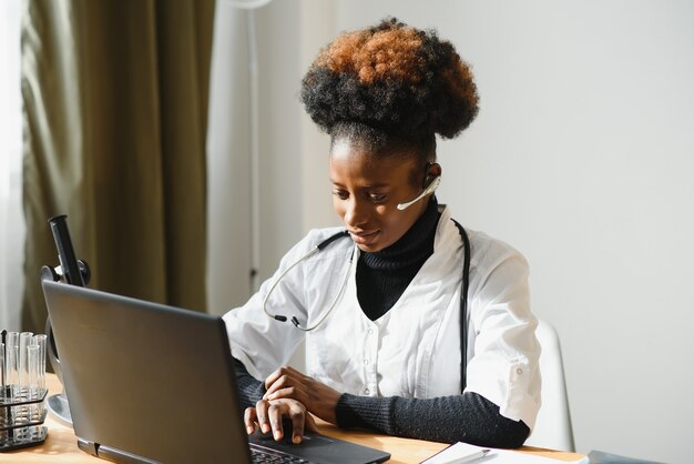 Sonriente doctora afroamericana gp usa bata médica blanca con computadora portátil en el lugar de trabajo.