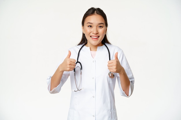 Sonriente doctora asiática, vestida con bata médica, mostrando los pulgares hacia arriba complacidos, recomendando productos para la salud, de pie sobre fondo blanco.