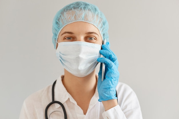 Foto sonriente doctora positiva vestida con uniforme médico, gorra y mascarilla quirúrgica hablando por teléfono con el paciente, mirando a la cámara con expresión facial optimista.