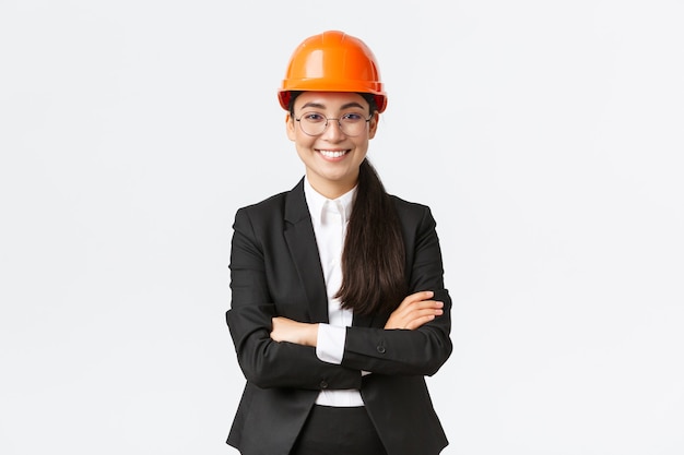 Sonriente empresaria asiática profesional en casco de seguridad y traje de brazos cruzados y mirando confiada, vendiendo nuevos edificios. Gerente de construcción, ingeniero mostrando, saludando a los inversores