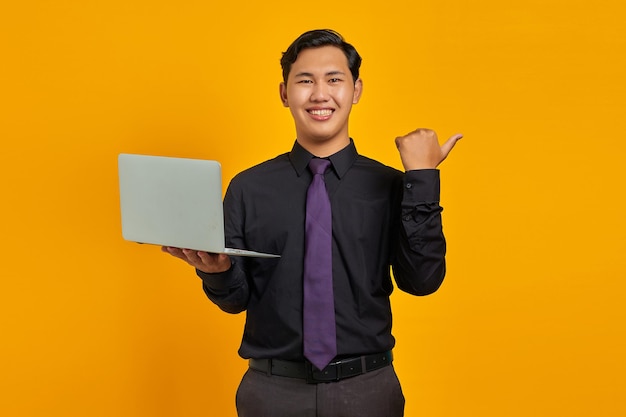 Sonriente empresario asiático sosteniendo portátil mientras apunta con el pulgar al espacio de copia sobre fondo amarillo