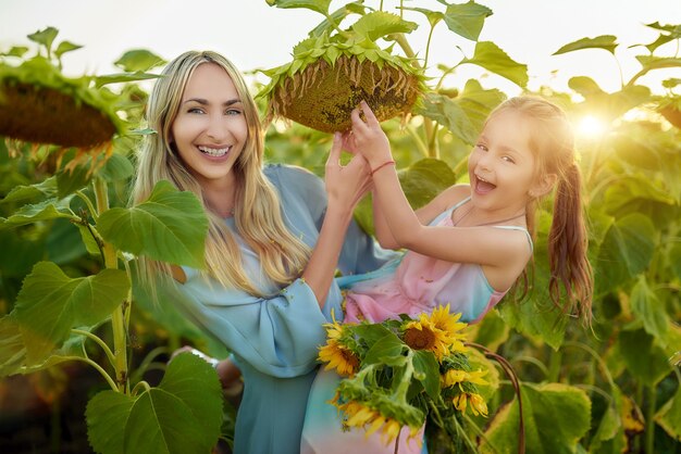 Sonriente y encantadora madre e hija cepillan un gran girasol para llegar a las semillas mientras caminan por el campo en un cálido día de verano. Concepto de fin de semana familiar