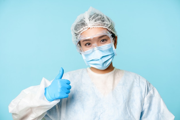 Sonriente enfermera o médico asiático, vistiendo ppe, equipo de protección personal, mostrando los pulgares hacia arriba, recomendando la vacunación, fondo azul