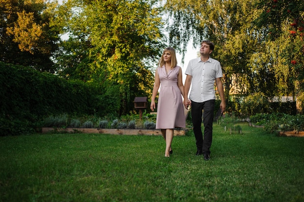 Sonriente esposo y esposa caminando sobre la hierba tomados de la mano en el parque verde debajo de las ramas de los árboles