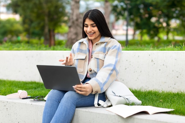 Sonriente estudiante hispana haciendo videollamadas en una laptop al aire libre usando una computadora para teleconferencia