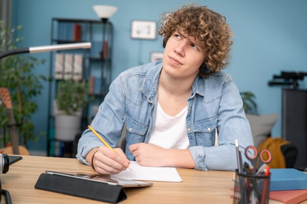 Sonriente estudiante milenario americano con auriculares sentado en el escritorio pensando en tomar notas