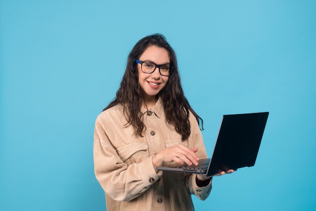 Sonriente estudiante morena milenaria europea en gafas escribiendo en la computadora portátil aislada en azul