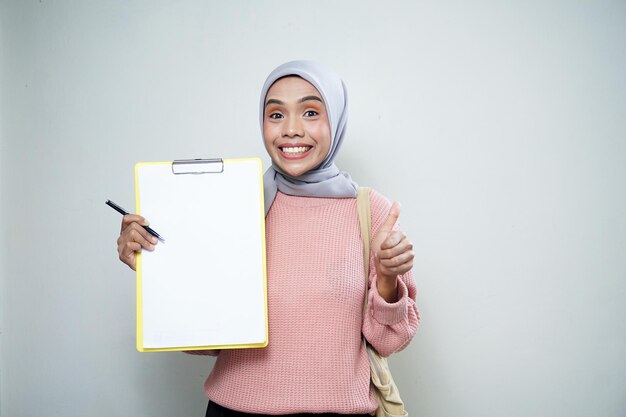 Sonriente estudiante musulmana asiática en suéter rosa con bolsa sosteniendo tablero y señalando tablero vacío