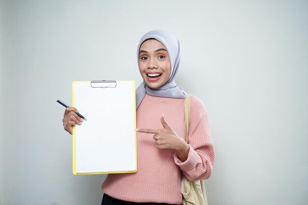 Sonriente estudiante musulmana asiática en suéter rosa con bolsa sosteniendo tablero y señalando tablero vacío