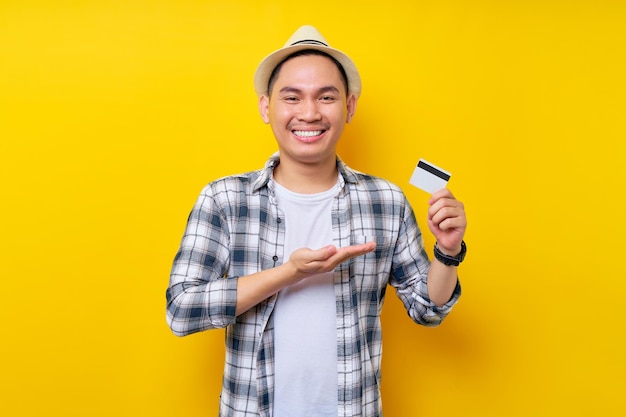 Sonriente feliz etnia asiática joven de 20 años con ropa informal que muestra una tarjeta de crédito en la mano que presenta un producto aislado en un retrato de estudio de fondo amarillo Concepto de estilo de vida de la gente