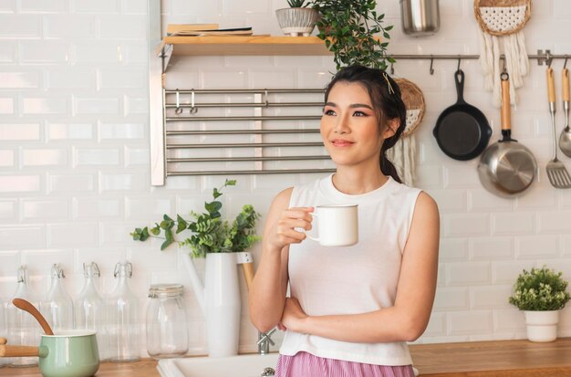 Sonriente feliz hermosa mujer asiática relajándose bebiendo una taza de café caliente o té en la cocina en casa