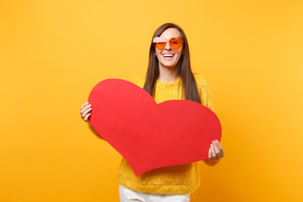 Sonriente a feliz mujer joven en suéter de piel y anteojos de corazón naranja con corazón rojo en blanco vacío aislado sobre fondo amarillo brillante. Personas sinceras emociones, concepto de estilo de vida. Área de publicidad.