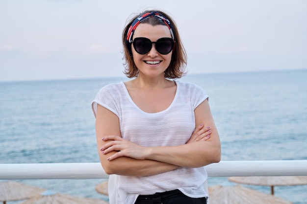 Sonriente feliz mujer madura confiada en gafas de sol mirando a la cámara con los brazos cruzados, fondo de noche de verano de cielo de mar