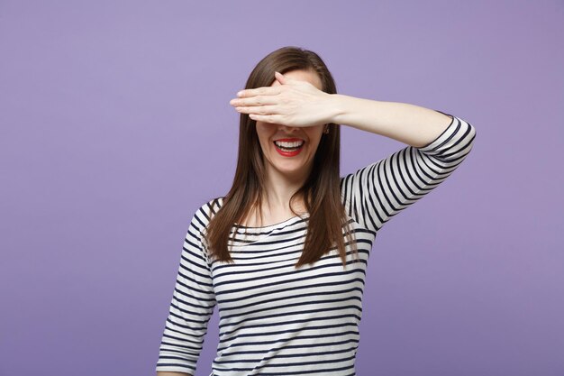 Sonriente y graciosa joven morena con ropa informal a rayas posando aislada en un retrato de estudio de fondo morado violeta. Concepto de estilo de vida de las personas. Simulacros de espacio de copia. Cubriéndose los ojos con la mano.