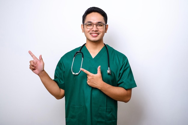 Sonriente y guapo enfermero médico asiático con batas y estetoscopio señalando con el dedo la esquina superior izquierda que muestra el banner de la clínica en línea