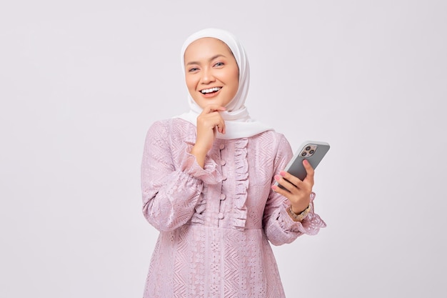 Sonriente y hermosa joven musulmana asiática con hiyab y vestido morado sosteniendo un teléfono móvil mientras toca la barbilla y piensa en algo aislado en el fondo del estudio blanco