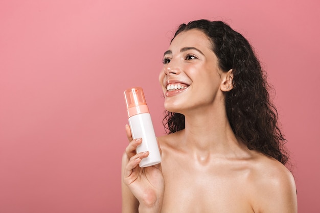 Sonriente a hermosa mujer joven posando aislada sobre pared rosa cuidar su piel con cosméticos.