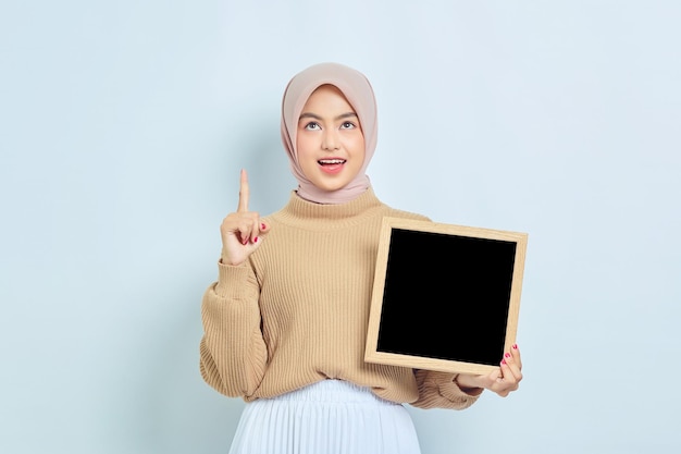 Sonriente hermosa mujer musulmana asiática en suéter marrón mostrando pizarra y señalando con el dedo hacia arriba aislado sobre fondo blanco Concepto de estilo de vida religioso de la gente