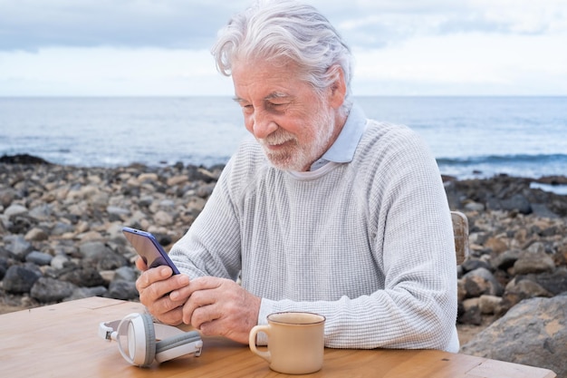 Sonriente hermoso anciano sosteniendo un teléfono móvil leyendo un mensaje al aire libre sentado en la playa Atractivo anciano nómada de pelo blanco mirando el teléfono inteligente